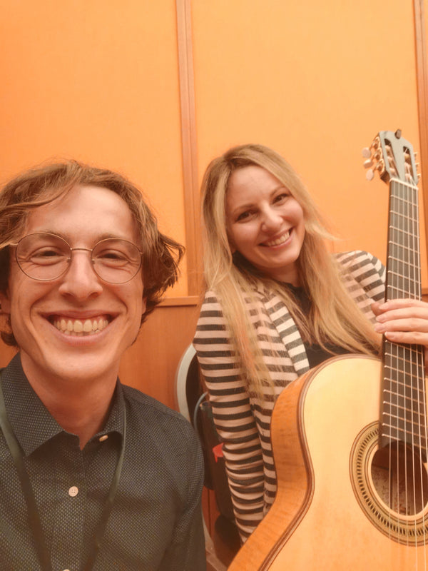 Two people smiling, one holding a guitar, against an orange wall.