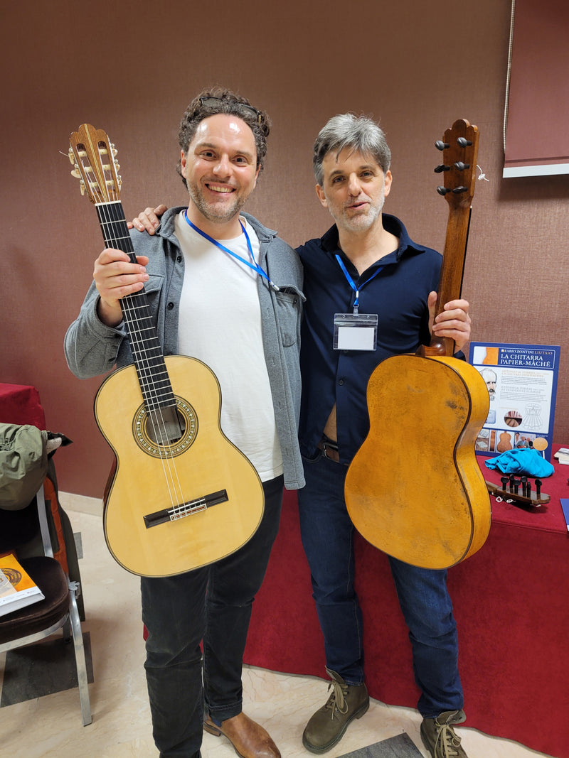 Two people holding classical guitars indoors with a red carpet and wall in the background.