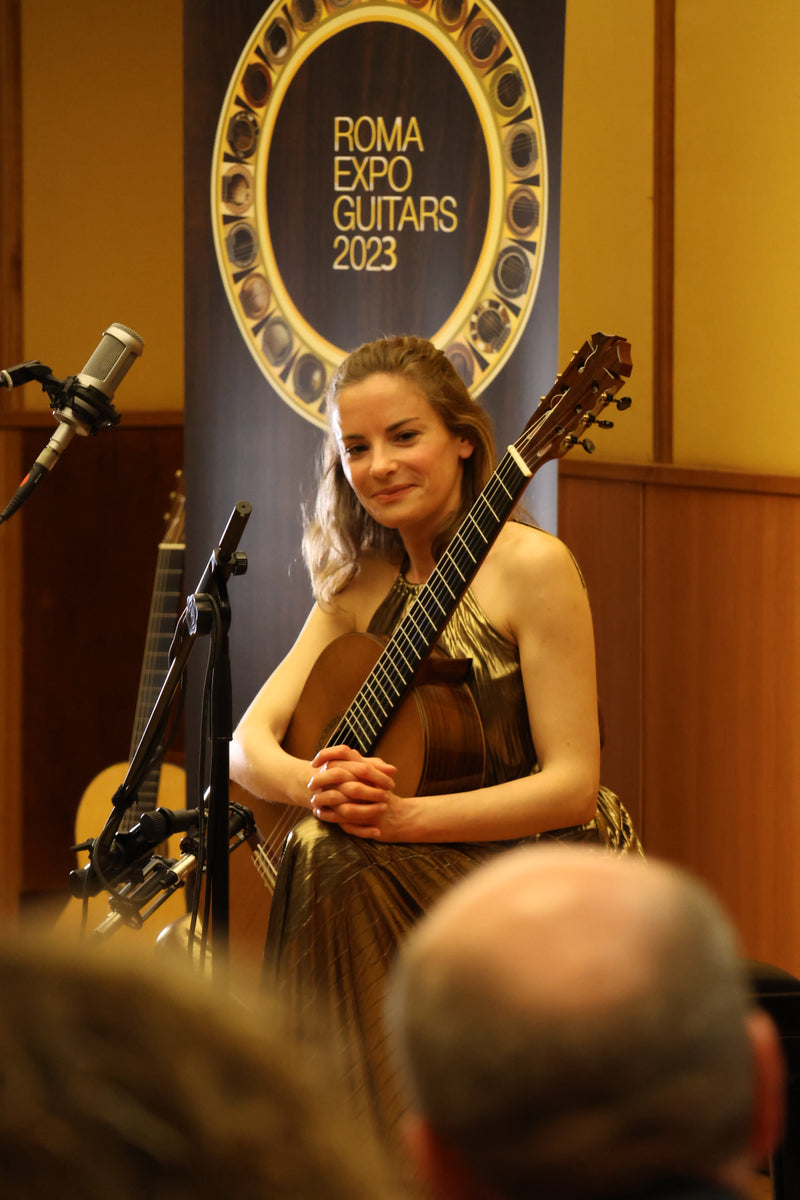 Ana Vidovic playing a guitar with 'Roma Expo Guitars' logo in the background