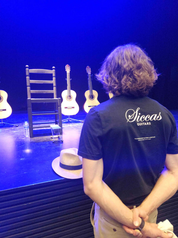 Person wearing a 'Siccas Guitars' shirt standing in front of guitars on a stage with blue lighting.