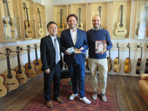 Three men standing in a guitar store holding books, with guitars displayed on the wall behind them.