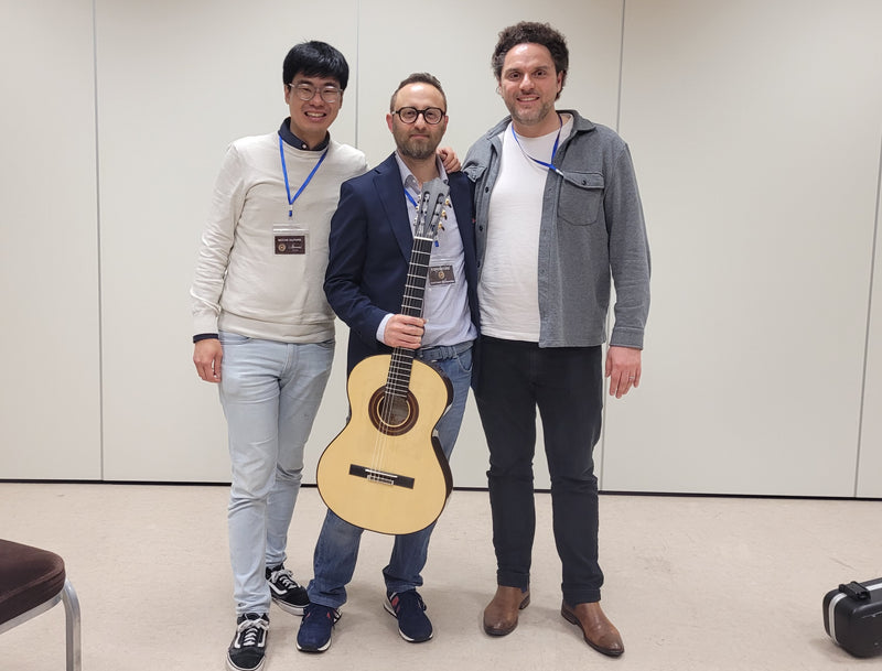 Three men standing together, with one holding a guitar, in an indoor setting.