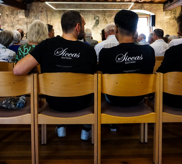 Two people wearing 'Siccas Guitars' shirts sitting in a room with wooden chairs.