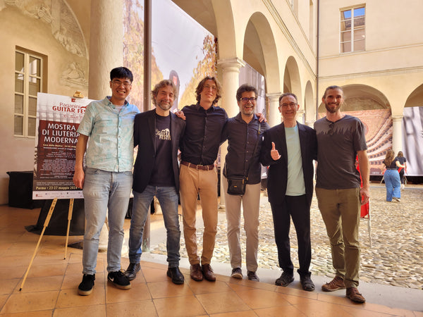 Group of six men posing for a photo in an outdoor setting with architectural columns and posters in the background.