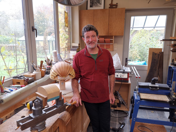 Man in a workshop holding a wooden object, surrounded by tools and equipment.